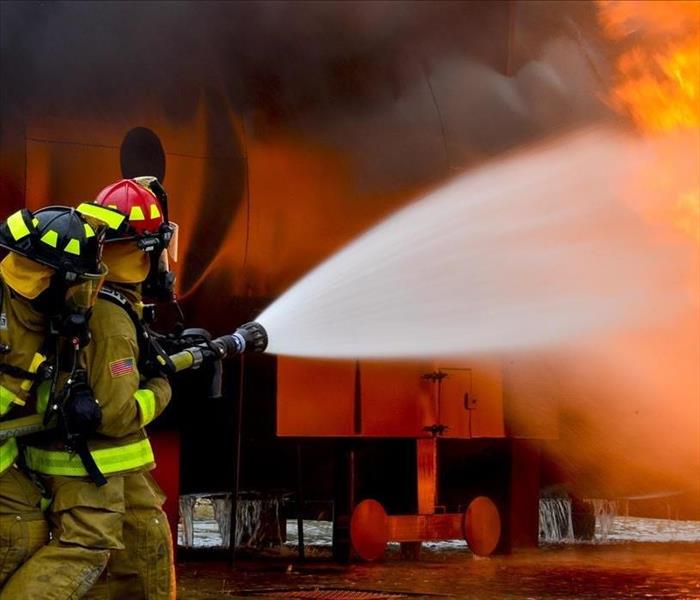 Two fire fighters hold a fire hose, aiming at large flames.